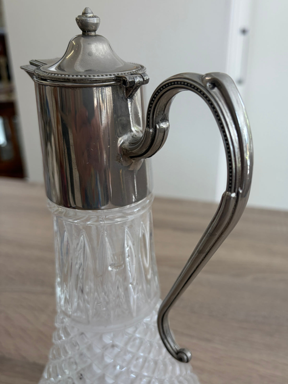 A vintage silver-toned wine decanter
with a lid and curved handle, mounted on a clear cut-crystal glass base, shown on a wooden tabletop.