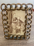 A rectangle antique-style picture frame with an ornate interlocking chain-like border in a warm metallic gold/bronze tone, holding an old sepia-toned portrait photograph of a couple, displayed on a wooden surface.