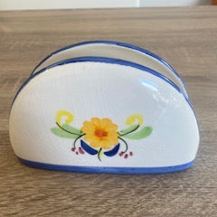 A white ceramic napkin holder with blue trim and a yellow floral design, resting on a wooden tabletop.