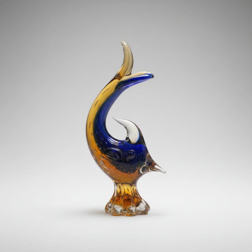 A decorative glass shaped like a leaping  fish, featuring a curved body in amber yellow, dark blue, and black, with a translucent orange tail, mounted on a white tiled wall.