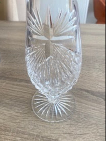 A clear cut-crystal water font with a religious cross and starburst pattern on the bowl and stem, resting on a light brown wooden tabletop.