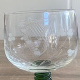 A clear glass goblet with an etched harp  design on the bowl, supported by a dark green ribbed stem and base, set on a wooden tabletop. 